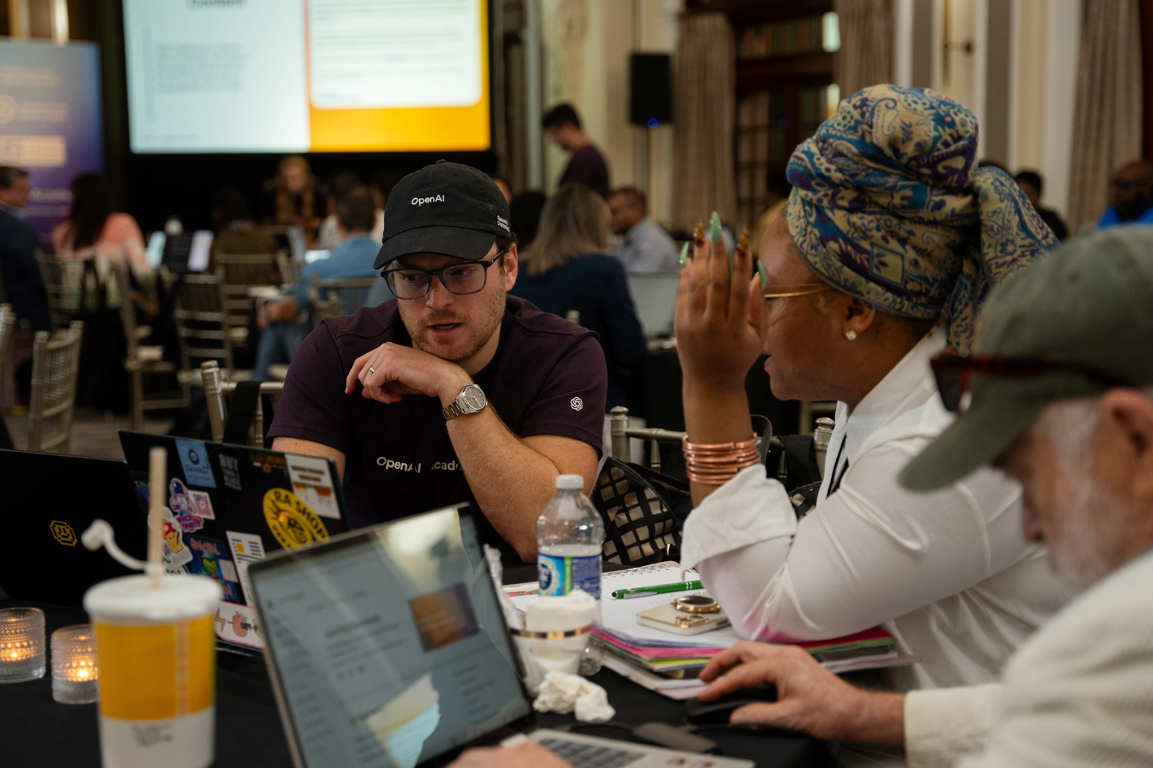 An OpenAI Academy team member talks with participants seated with laptops during a workshop.